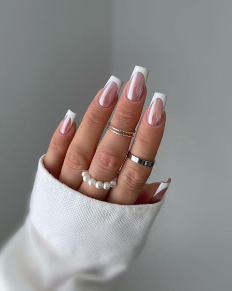 Unhas quadradas com francesinha branca e base rosada, numa mão com anéis prateados e de pérolas.