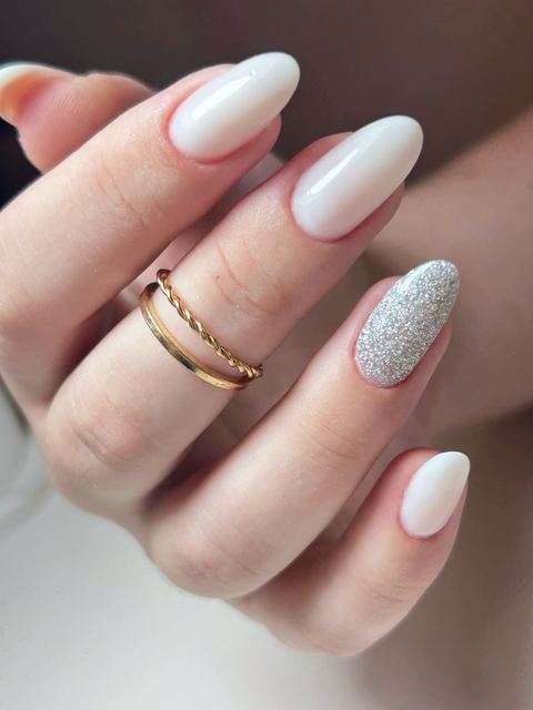 Unhas amendoadas em branco leitoso, uma delas com glitter prata, e anéis dourados.